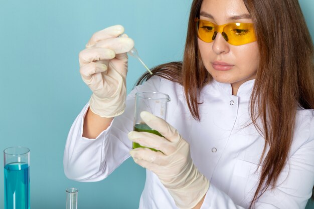 Front view of young female chemist in white suit in front of table working with different solutions