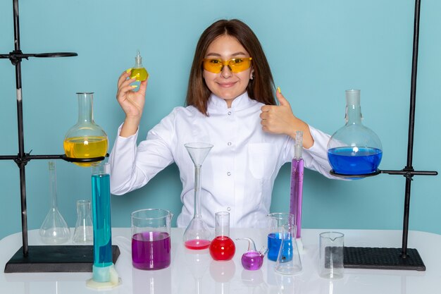 ソリューションの操作と笑顔のテーブルの前に白いスーツの若い女性化学者の正面図