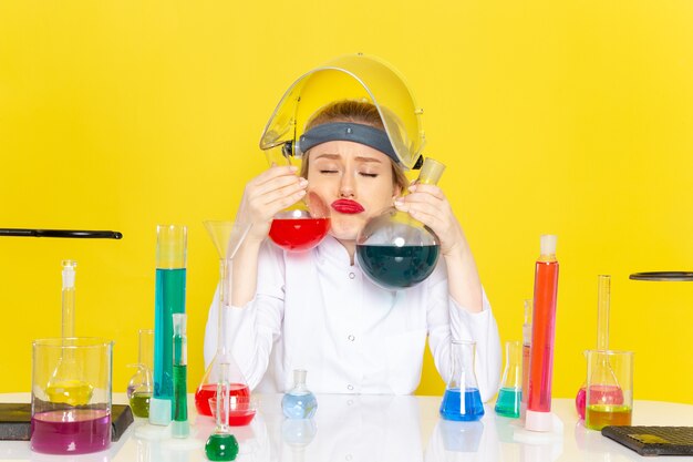 Front view young female chemist in white suit sitting and holding different solutions on the yellow space chemistry science s process job