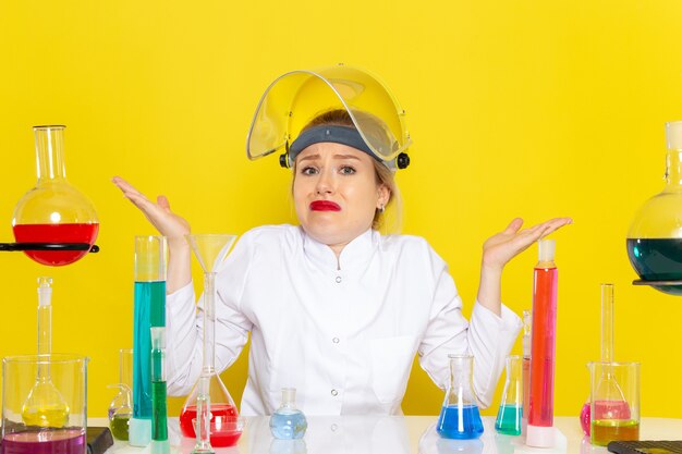 Front view young female chemist in white suit sitting with different solutions with confused expression on the yellow space chemistry science s