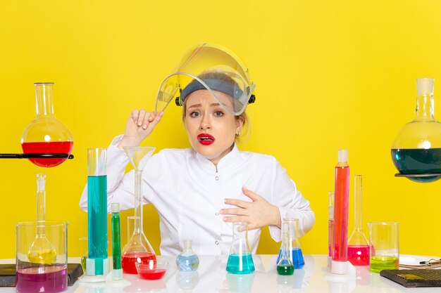 Front view young female chemist in white suit with ed solutions working with them wearing helmet on the yellow space chemistry  job