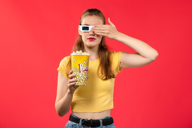 Вид спереди молодая женщина в кинотеатре, держащая попкорн в солнцезащитных очках -d на светло-красной стене, кинотеатр, закуска, веселый фильм
