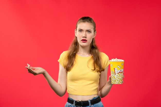 Вид спереди молодая женщина в кинотеатре, держащая пакет попкорна и смущенная на светло-красной стене, кинотеатр, кинотеатр, забавный фильм