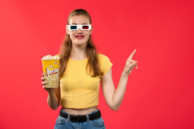 Вид спереди молодая женщина в кино с пакетом попкорна в солнцезащитных очках -d на светло-красной стене кино театр кинотеатр забавный фильм
