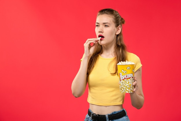 Вид спереди молодая женщина в кинотеатре держит пакет попкорна и ест на красной стене.