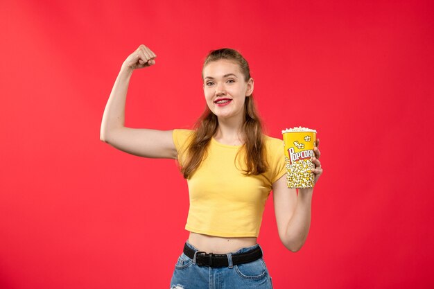 Front view young female at cinema holding popcorn package and flexing on red wall movies theater cinema snack female fun film