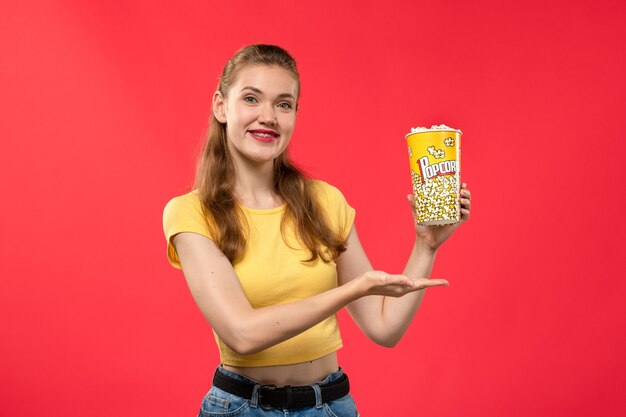 Вид спереди молодая женщина в кино, держащая пакет попкорна с легкой улыбкой на красной стене, кино, кинотеатр, женский забавный фильм