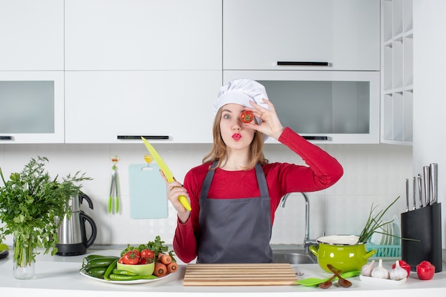 Front view young female cook in apron holding up tomato in front of her eye
