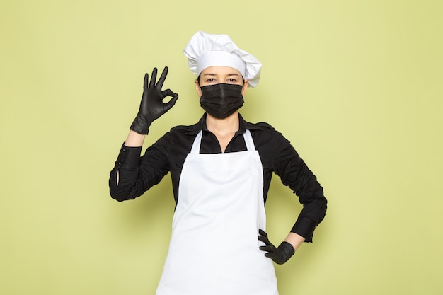 Free photo a front view young female cook in black shirt white cook cape white cap posing in black gloves black mask posing