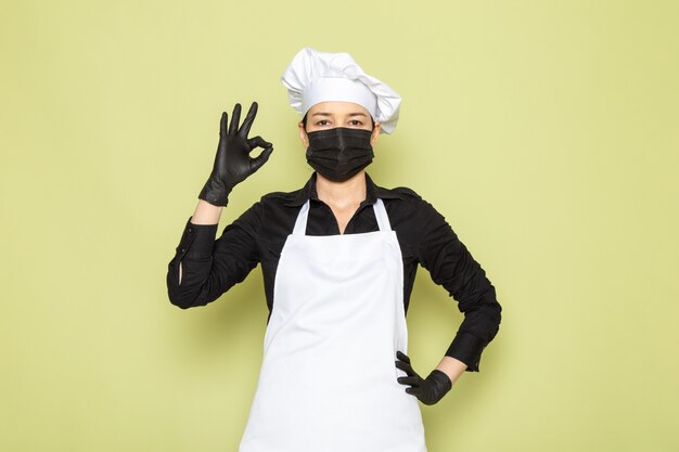 A front view young female cook in black shirt white cook cape white cap posing in black gloves black mask posing