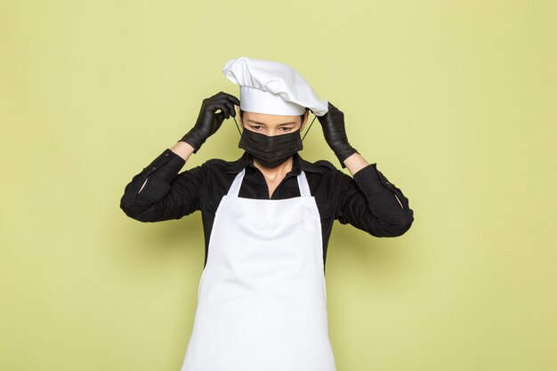 Free photo a front view young female cook in black shirt white cook cape white cap posing in black gloves wearing black mask posing