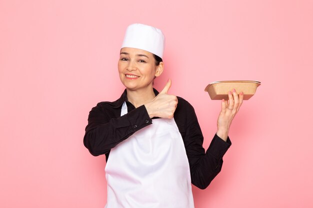 A front view young female cook in black shirt white cook cape white cap posing holding milk brown bowl smiling