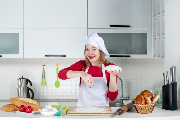 Вид спереди молодая женщина-повар в поварской шляпе и фартуке держит тесто, посыпая мукой на кухне