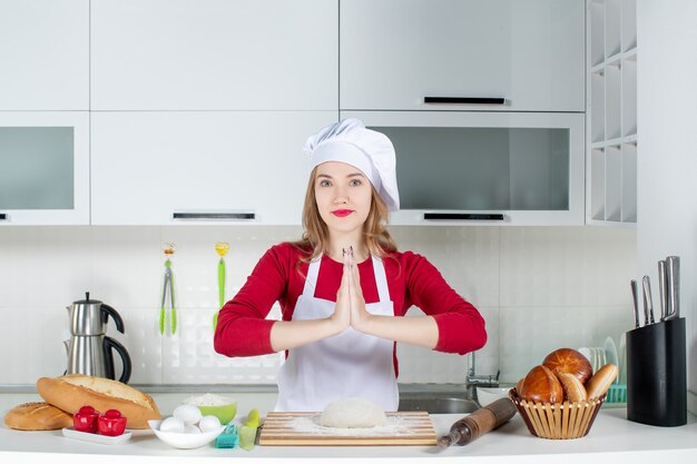 Front view young female cook in cook hat and apron joining her hands together in the kitchen