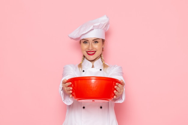 Бесплатное фото Вид спереди молодая женщина-повар в белом костюме повара держит красную пластиковую миску на розовом космическом поваре