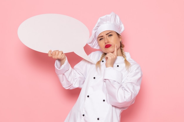Бесплатное фото Вид спереди молодая женщина-повар в белом костюме повара держит белый знак с мыслящим выражением на розовой фотографии космического повара