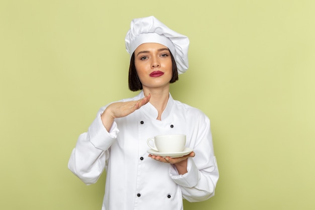 A front view young female cook in white cook suit and cap holding cup on the green wall