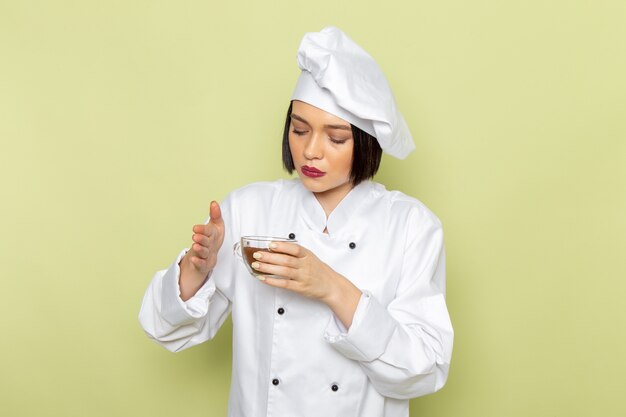 A front view young female cook in white cook suit and cap holding cup of powdered coffee on the green wall