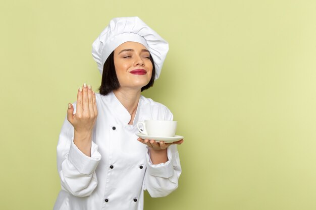 흰색 요리사 양복과 녹색 벽에 미소로 컵을 들고 모자에 전면보기 젊은 여성 요리사 레이디 작업 음식 요리 색상