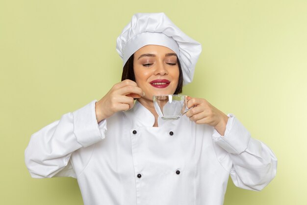 A front view young female cook in white cook suit and cap holding an empty cup on the green wall