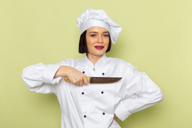 A front view young female cook in white cook suit and cap holding knife on the green wall lady work food cuisine color