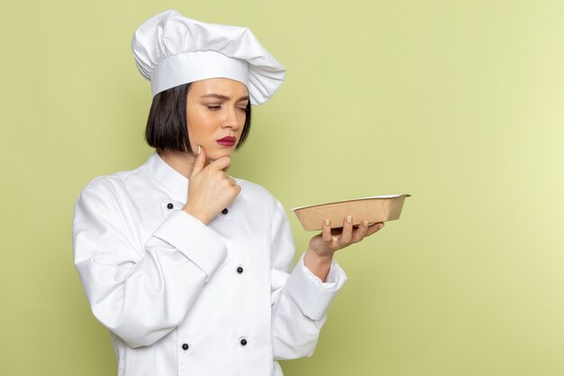 흰색 요리사 양복과 녹색 벽에 생각 식으로 패키지를 들고 모자에 전면보기 젊은 여성 요리사 레이디 작업 음식 요리 색상