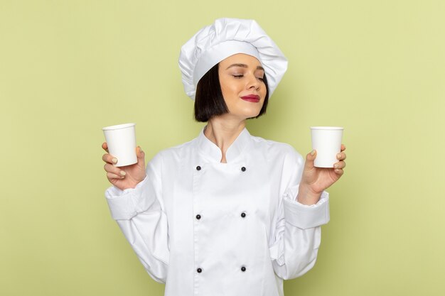 白いクックスーツと緑の壁にプラスチックカップを保持しているキャップで正面の若い女性クック女性の仕事食品料理色