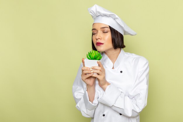 A front view young female cook in white cook suit and cap holding and smelling green plant on the green wall