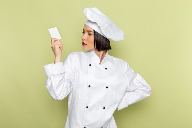 흰색 쿡 양복과 녹색 벽에 흰색 카드를 들고 모자에 전면보기 젊은 여성 요리사 레이디 작업 음식 요리 색상