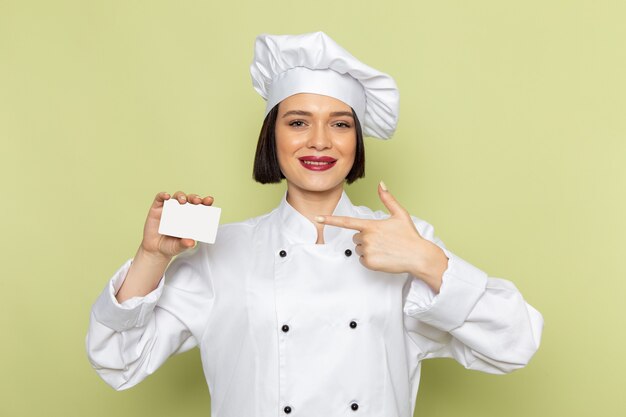 A front view young female cook in white cook suit and cap holding white card with smile on the green wall lady work food cuisine color