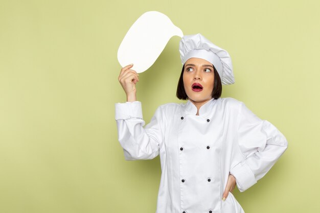 A front view young female cook in white cook suit and cap holding white sign on the green wall lady work food cuisine color