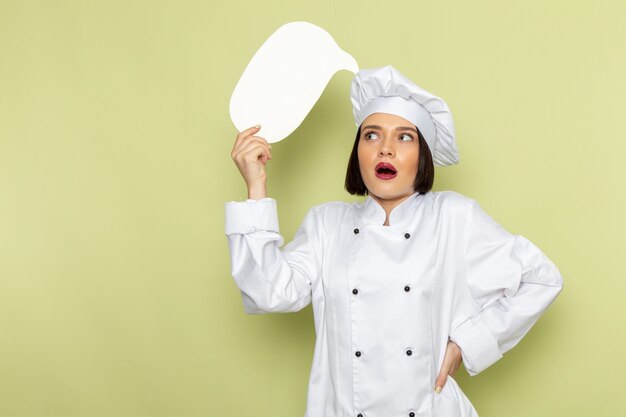 흰색 요리사 양복과 녹색 벽에 흰색 기호를 들고 모자에 전면보기 젊은 여성 요리사 레이디 작업 음식 요리 색상