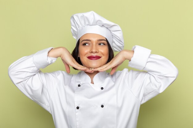 A front view young female cook in white cook suit and cap posing in alluring way on the green wall lady work food cuisine color