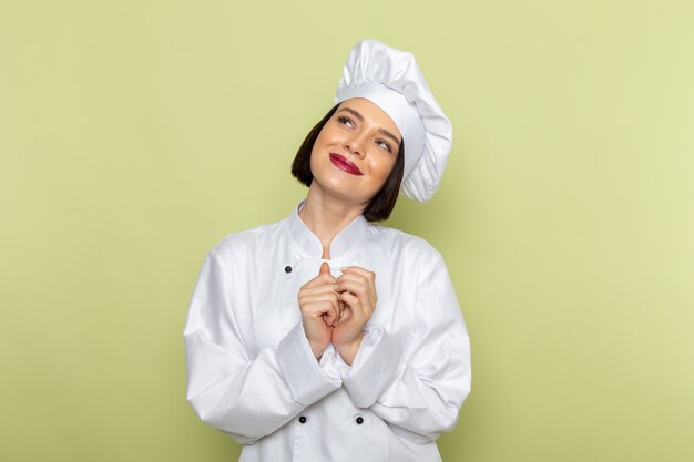 A front view young female cook in white cook suit and cap posing and smiling on the green wall lady work food cuisine color