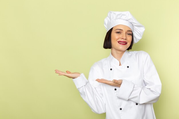 A front view young female cook in white cook suit and cap posing with smile on the green wall lady work food cuisine color