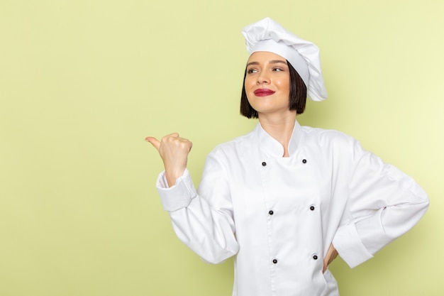 A front view young female cook in white cook suit and cap posing with smile on the green wall lady work food cuisine color