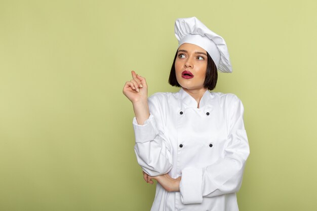 Un giovane cuoco femminile di vista frontale in vestito e cappuccio bianchi del cuoco che posano con l'espressione sorpresa sul colore di cucina dell'alimento del lavoro della signora della parete verde