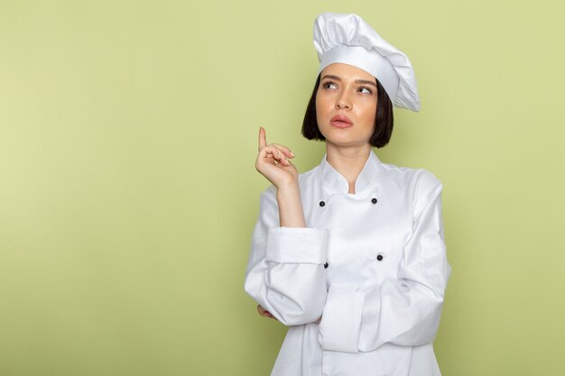 흰색 요리사 양복과 모자에 전면보기 젊은 여성 요리사 녹색 벽에 포즈 생각에 여자 일 음식 요리 색상