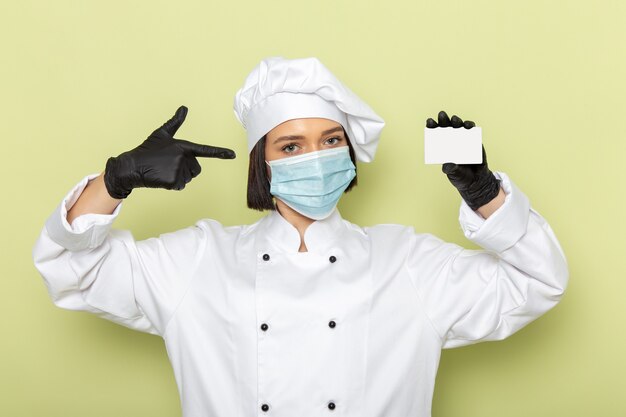 A front view young female cook in white cook suit and cap wearing gloves and mask on the green wall lady work food cuisine color
