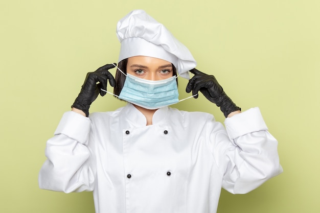 緑の壁の女性作業食品の色に白いクックスーツとキャップと手袋と滅菌マスクを身に着けている正面の若い女性クック