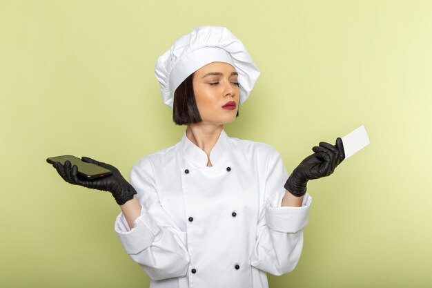 Free photo a front view young female cook in white cook suit and cap wearing gloves and sterile mask holding phone on the green wall lady work food color