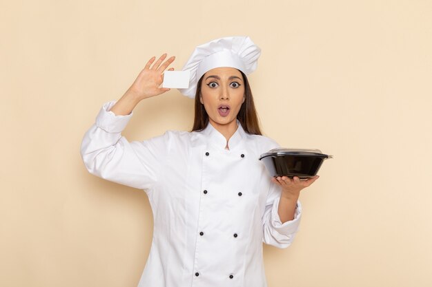明るい白い壁に黒いボウルとカードを保持している白いクックスーツの若い女性料理人の正面図
