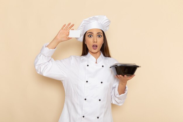 Front view of young female cook in white cook suit holding black bowl and card on light white wall