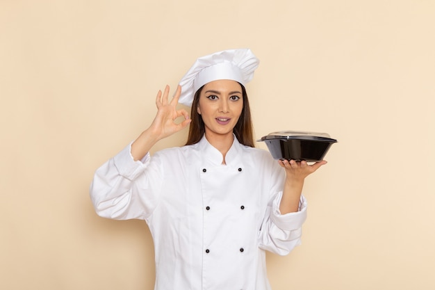 Front view of young female cook in white cook suit holding black bowl on light-white wall