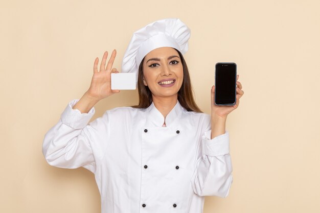 Front view of young female cook in white cook suit holding card and phone on light-white wall