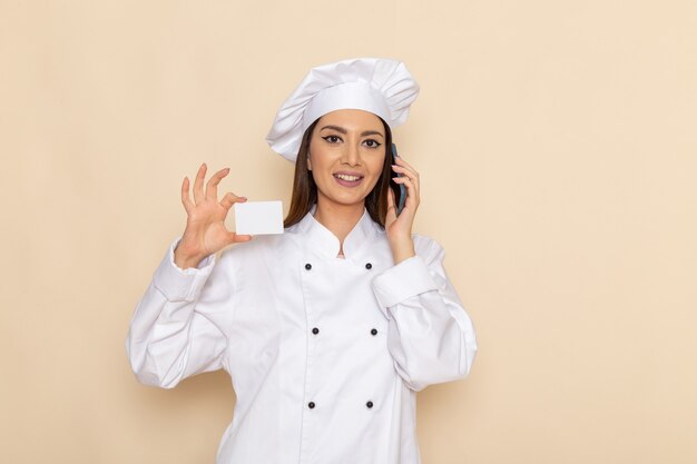 Front view of young female cook in white cook suit holding card and talking on the phone on light-white wall