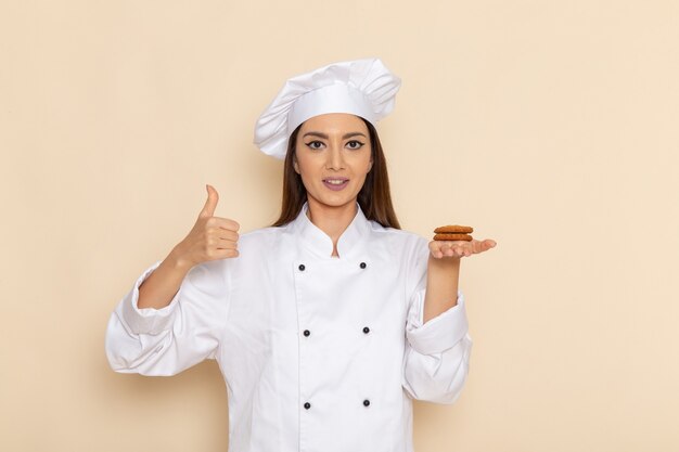 明るい白い壁にクッキーを保持している白いクックスーツの若い女性料理人の正面図