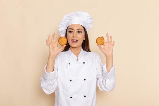 Front view of young female cook in white cook suit holding cookies on light-white wall