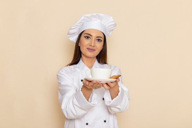 Front view of young female cook in white cook suit holding cup of coffee on light-white wall