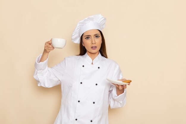 明るい白い壁にコーヒーカップを保持している白いクックスーツの若い女性料理人の正面図